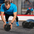 Man in blue shirt and black pants exercising with medicine balls in a gym setting.